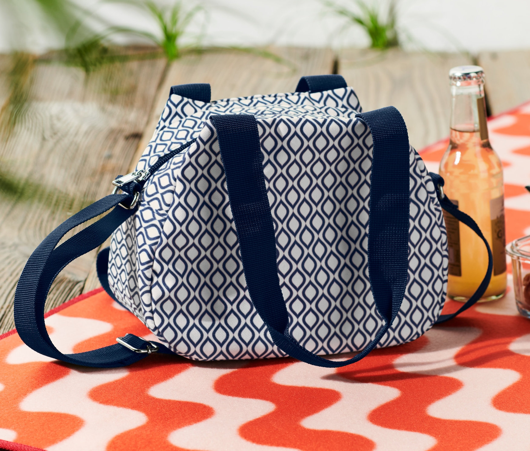 Niebiesko-biała mini torba termiczna leży obok napoju i miski z makaronem na pomarańczowo-białym, falistym ręczniku plażowym.