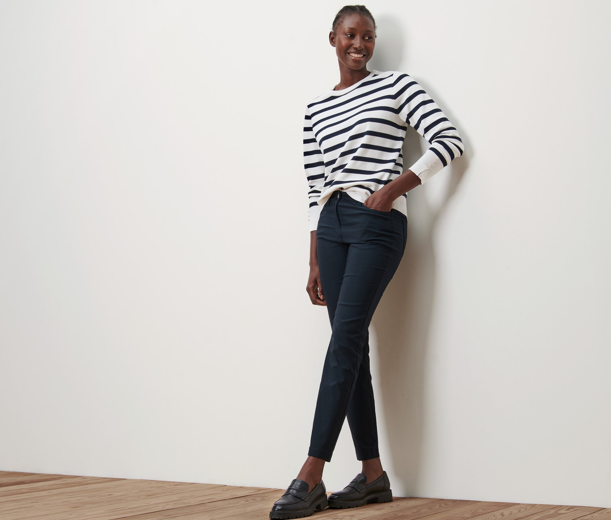 Kobieta opiera się o ścianę, ma na sobie granatowe spodnie z bengaliny, bluzkę w paski i czarne buty.