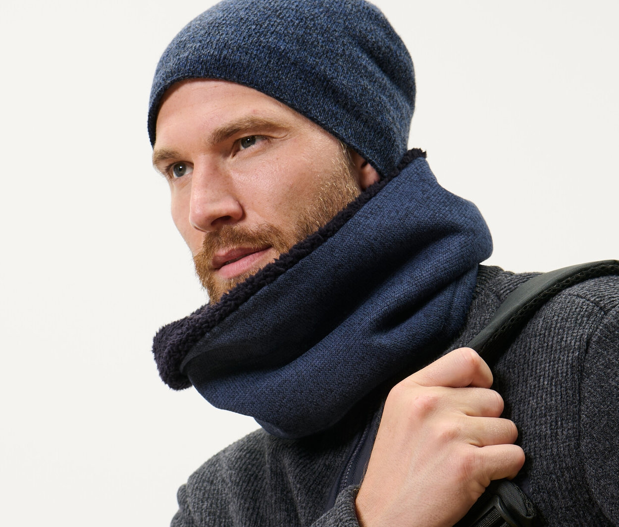 Mężczyzna z brodą ma na sobie granatowy szalik typu komin z dzianiny polarowej i czapkę oraz nosi plecak.