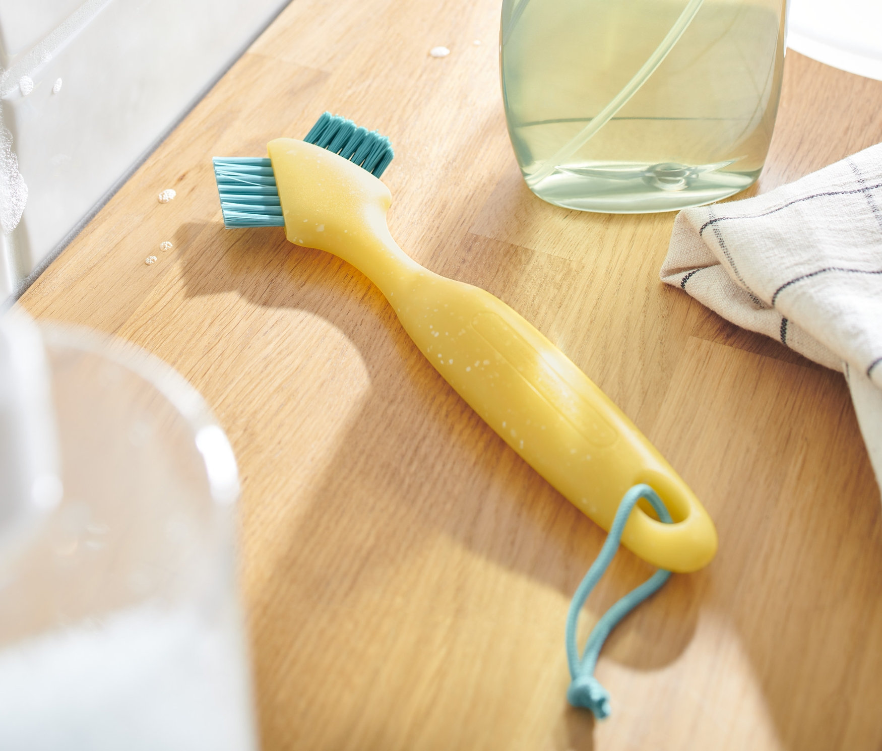 Żółta szczotka do czyszczenia fug z niebieskim włosiem leży na drewnianej powierzchni obok środka czyszczącego i ściereczki.