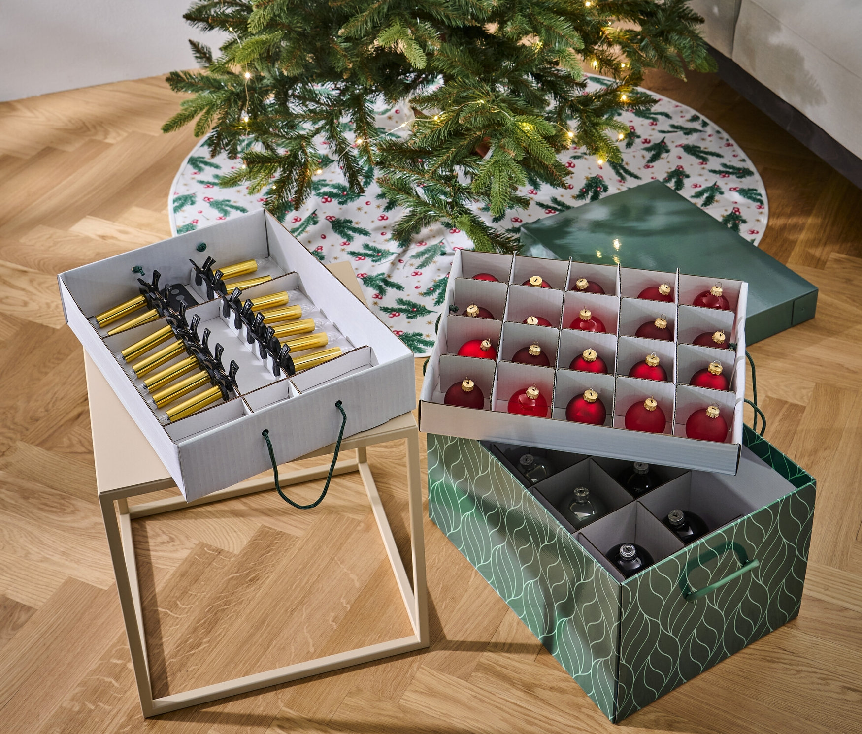 Choinka stoi obok dwóch pudełek na ozdoby świąteczne. Jedno pudełko jest białe i zawiera złote i czarne ozdoby, drugie jest zielone i zawiera czerwone i srebrne ozdoby.
