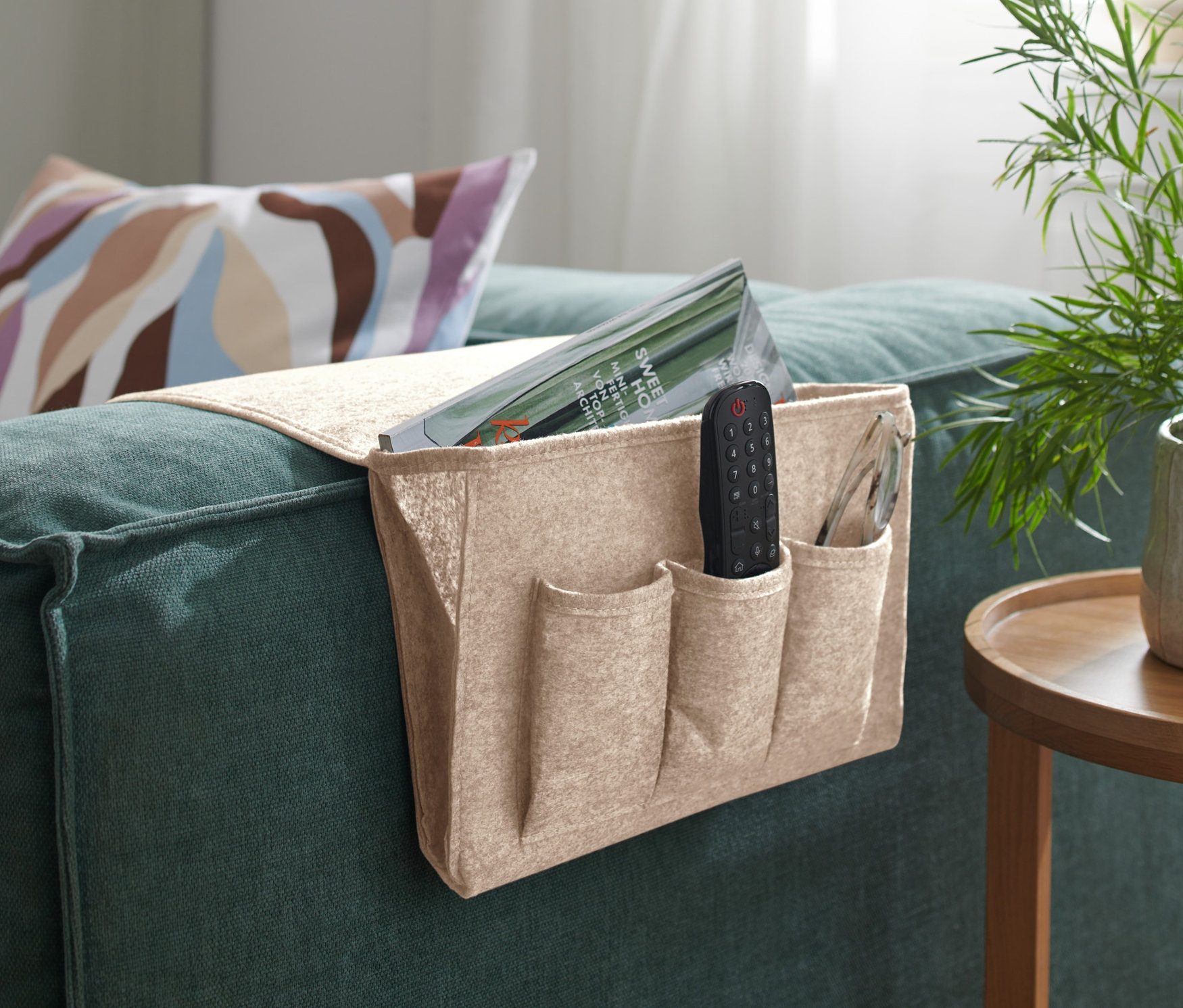 Beżowy organizer na łóżko lub sofę wisi na podłokietniku zielonej sofy, w środku znajduje się pilot i czasopismo.