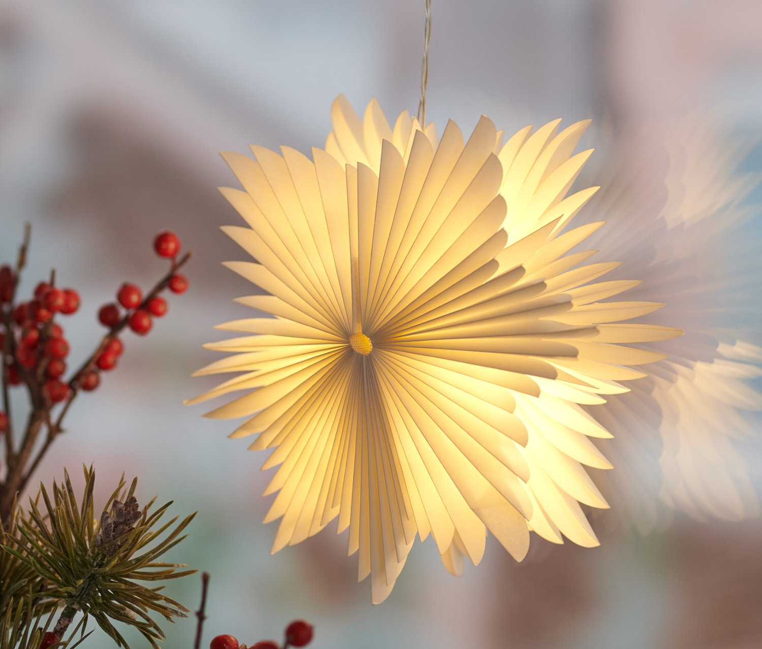 Mała papierowa gwiazda LED zawieszona przy oknie, obok gałęzi z czerwonymi jagodami.