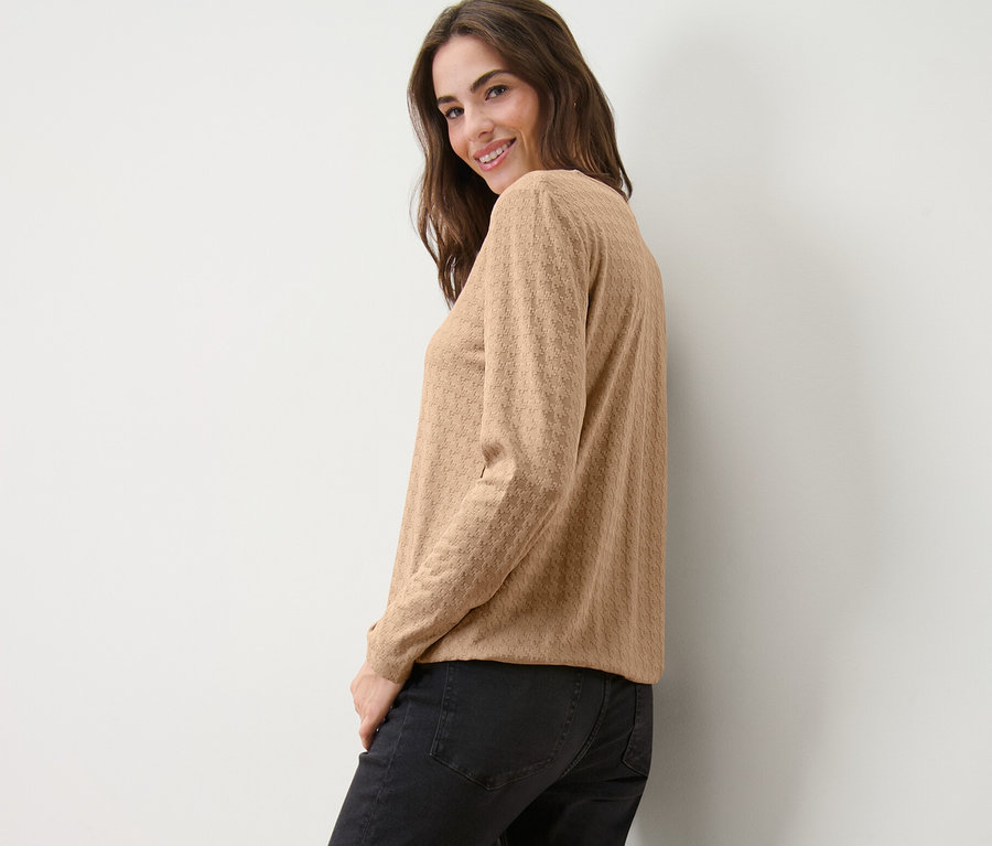 Uśmiechnięta kobieta spogląda przez ramię, pozując w karmelowej koszulce z długim rękawem ze strukturą i czarnych dżinsach.