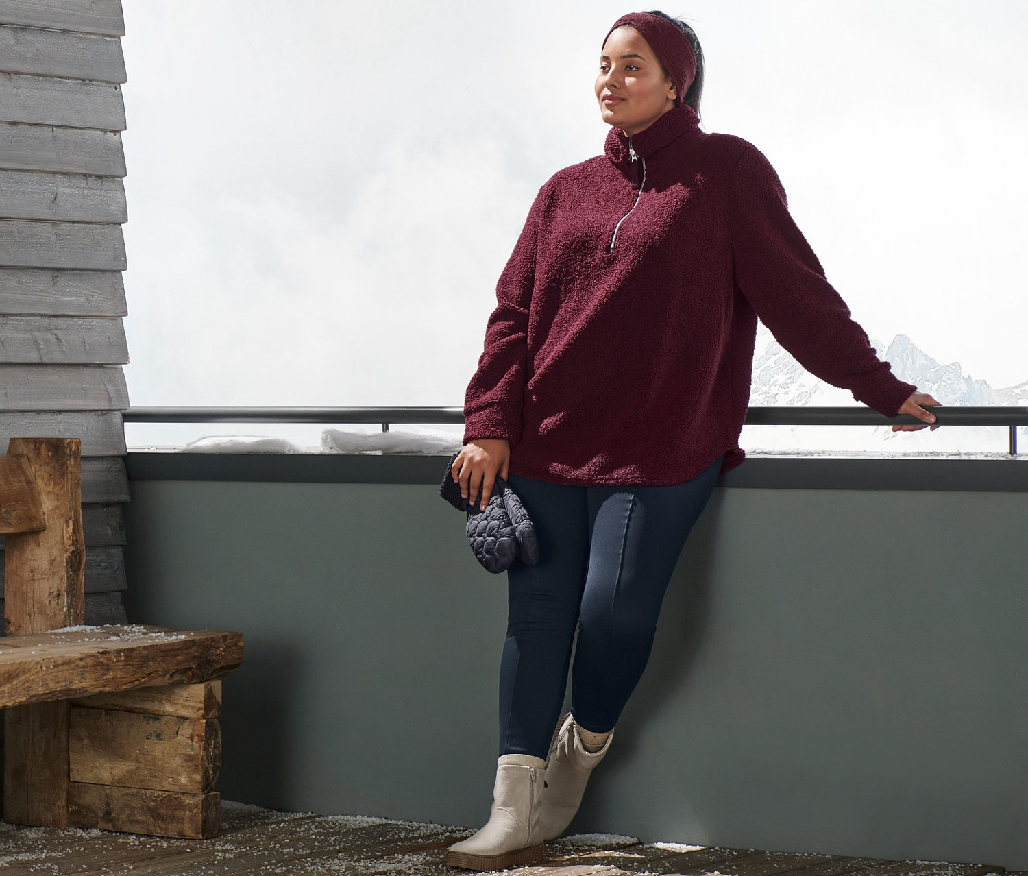 Kobieta opiera się o barierkę, ma na sobie bluzę pluszową oversize, bordową opaskę z dzianiny, niebieskie ocieplane legginsy i botki ze skóry zamszowej. W ręku trzyma pikowane rękawiczki z jednym palcem.