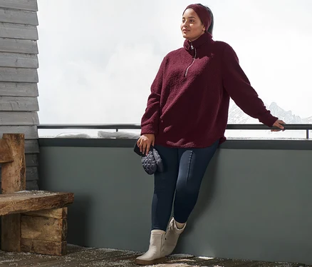Kobieta opiera się o barierkę, ma na sobie bluzę pluszową oversize, bordową opaskę z dzianiny, niebieskie ocieplane legginsy i botki ze skóry zamszowej. W ręku trzyma pikowane rękawiczki z jednym palcem.
