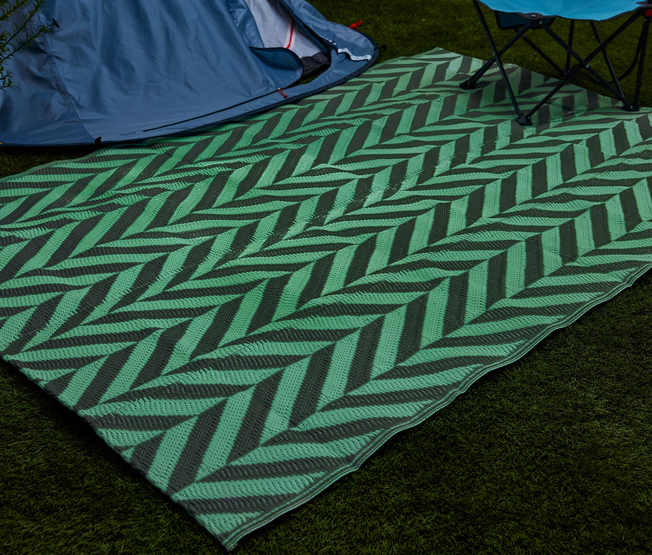 Zielony, fluorescencyjny dywan do namiotu ze wzorem szewronu leży na trawie obok namiotu i krzesła składanego.