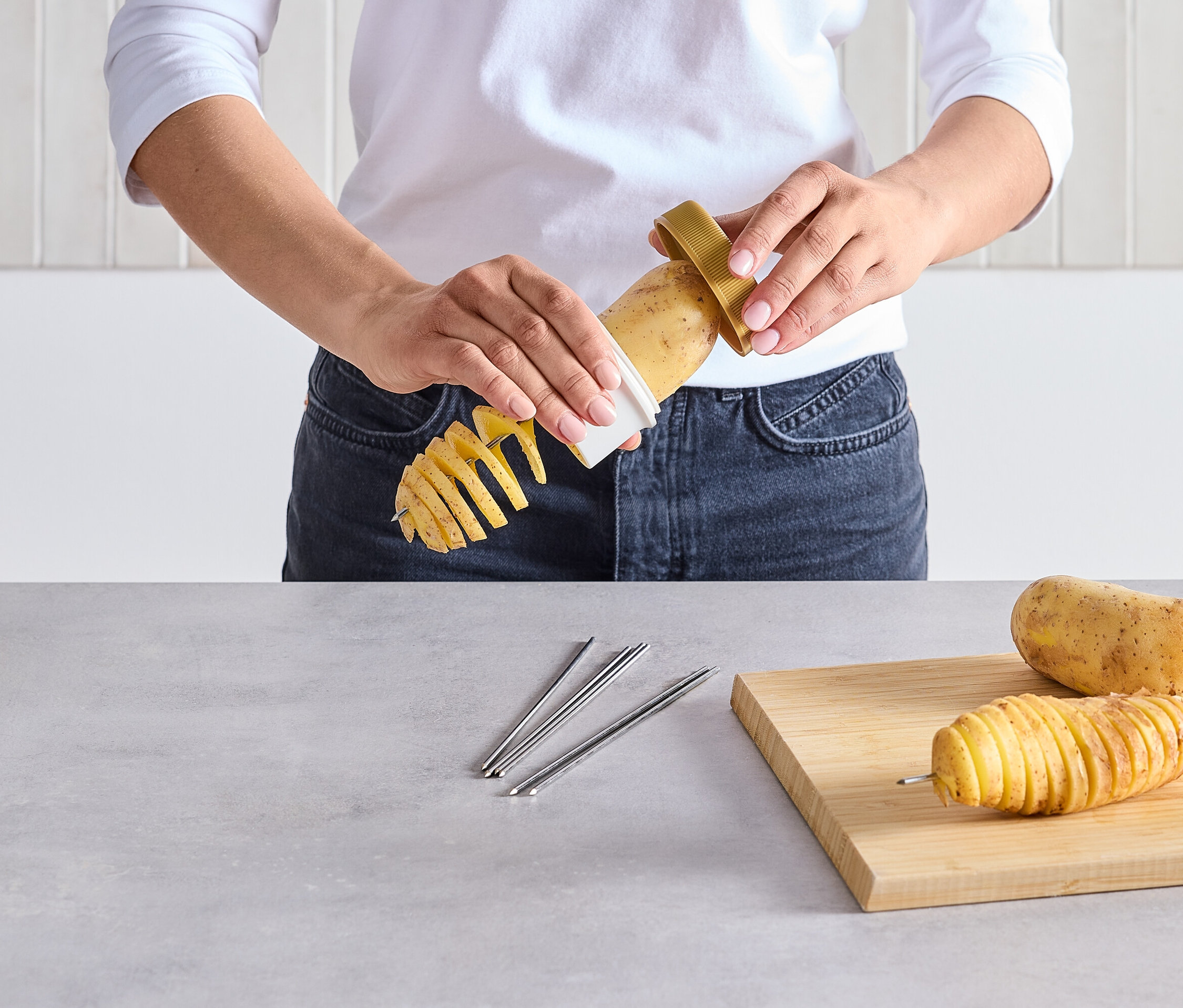 Osoba kroi ziemniaka na spiralę za pomocą specjalnego krajalnika. Na stole leżą pokrojony ziemniak na szpikulcu i surowy ziemniak.
