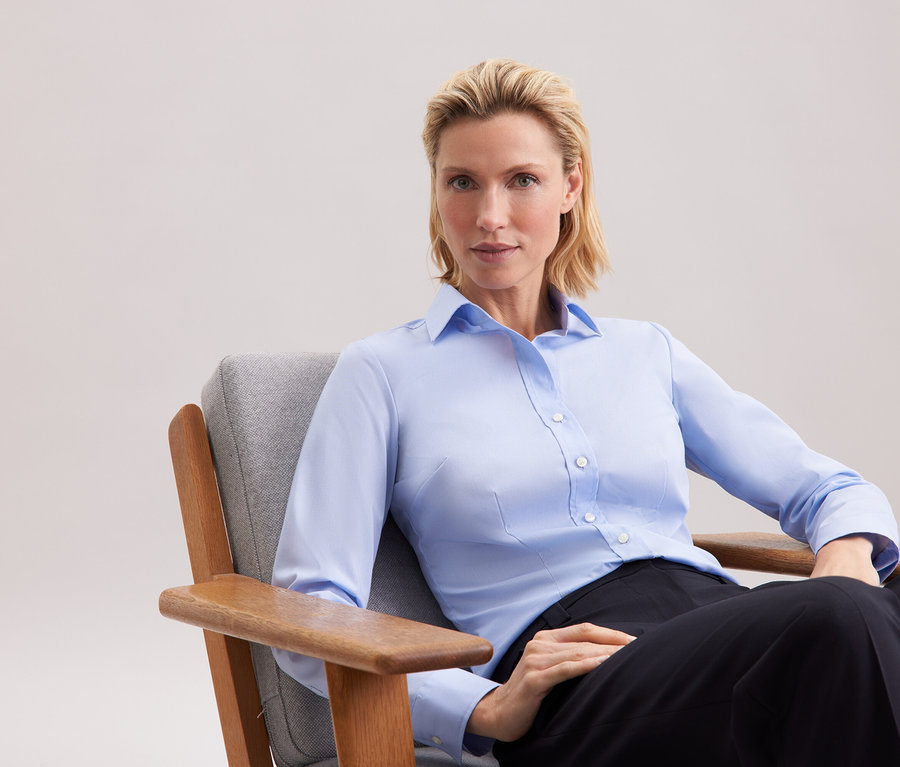 Blondynka siedzi w szarym fotelu, ubrana w niebieską koszulę i czarne spodnie.