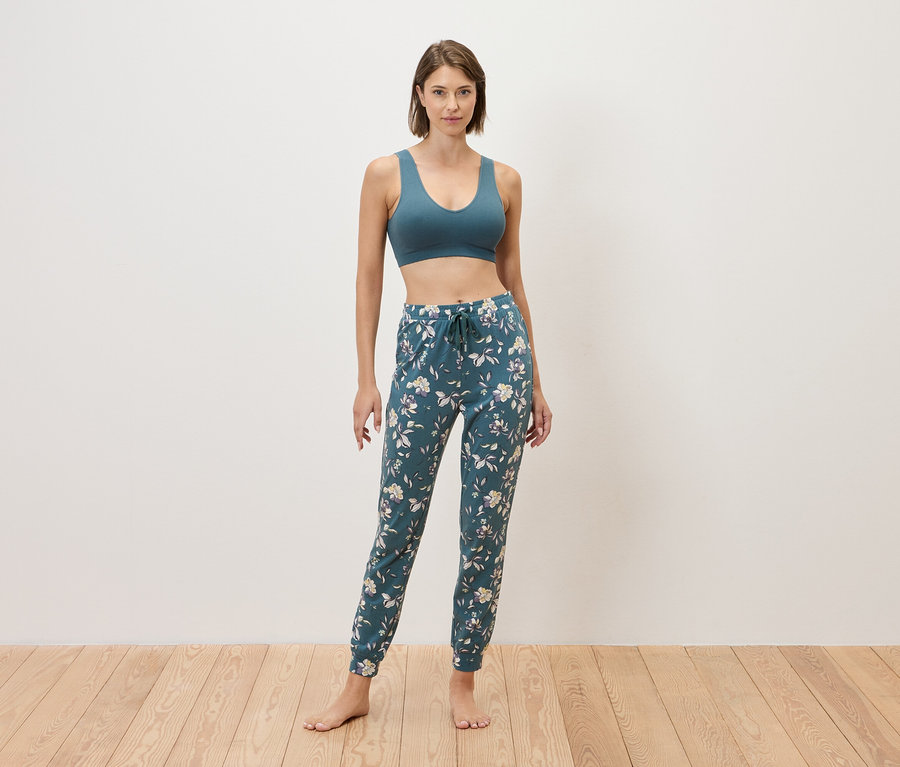 Kobieta stoi boso na tle ściany, ubrana w niebieski stanik sportowy i niebieskie spodnie dresowe w kwiaty.