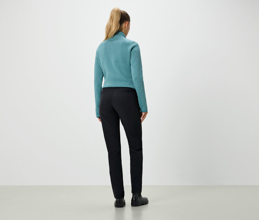 Widok z tyłu kobiety o blond włosach związanych w kucyk, ubranej w turkusowy sweter i czarne spodnie.