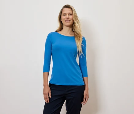Kobieta z blond włosami ma na sobie niebieską koszulkę z rękawem trzy czwarte i granatowe spodnie.