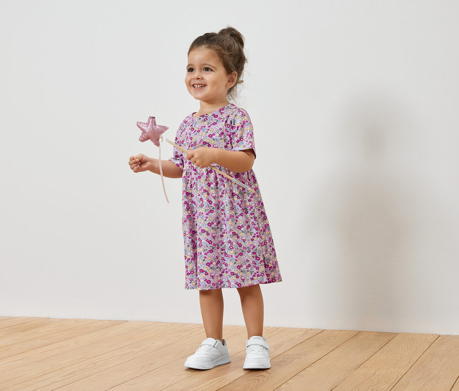 Uśmiechnięta dziewczynka trzyma gwiazdkę, ma na sobie kwiecistą sukienkę i tenisówki.