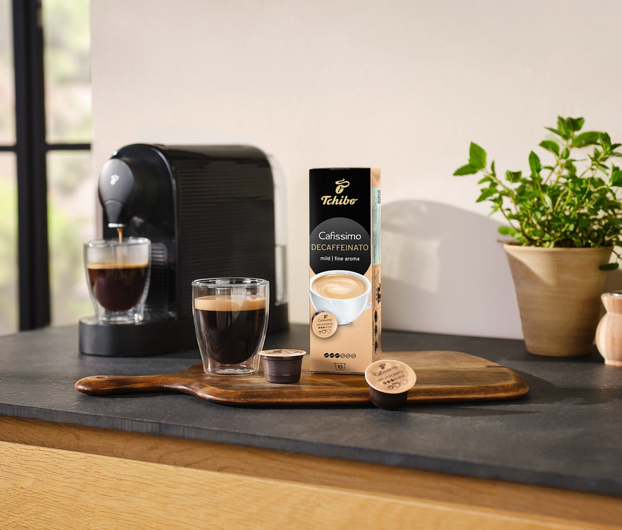 Ekspres do kawy nalewa kawę do szklanki obok opakowania kapsułek Cafissimo Decaffeinato.