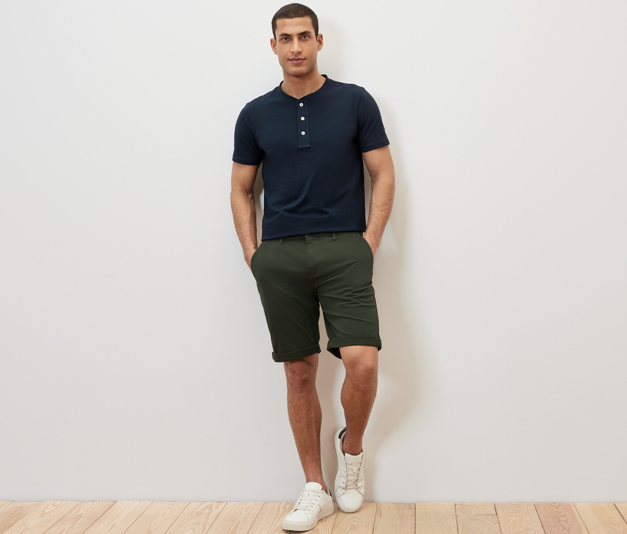 Mężczyzna stoi z rękami w kieszeniach szortów. Ma na sobie granatową koszulkę henley z krótkim rękawem.