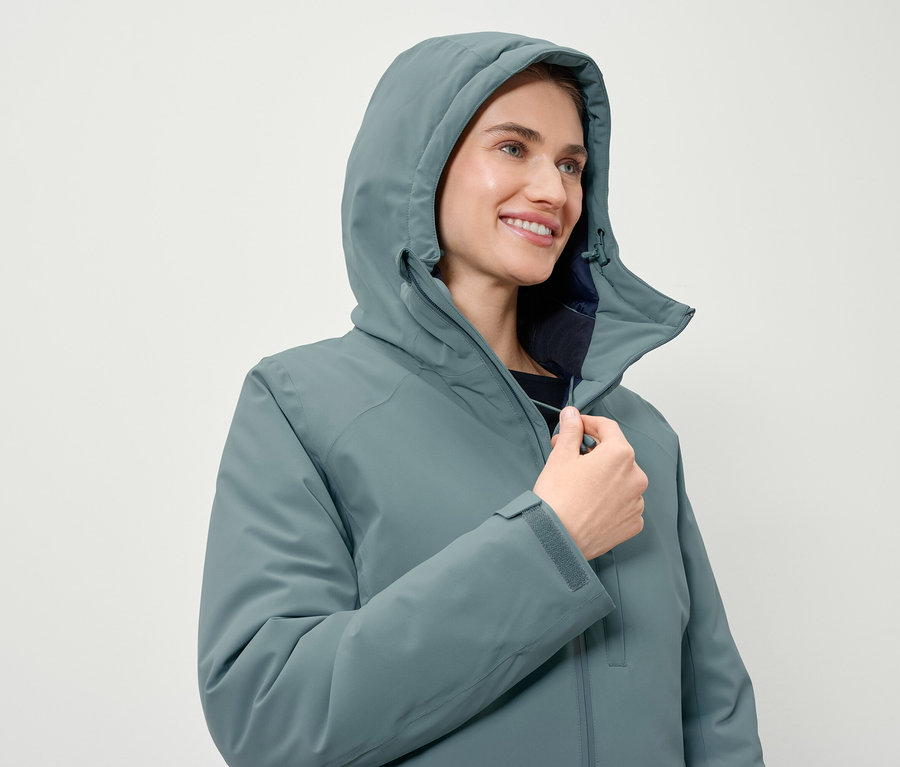Kobieta zapina na zamek kurtkę w kolorze niebiesko-zielonym z kapturem.