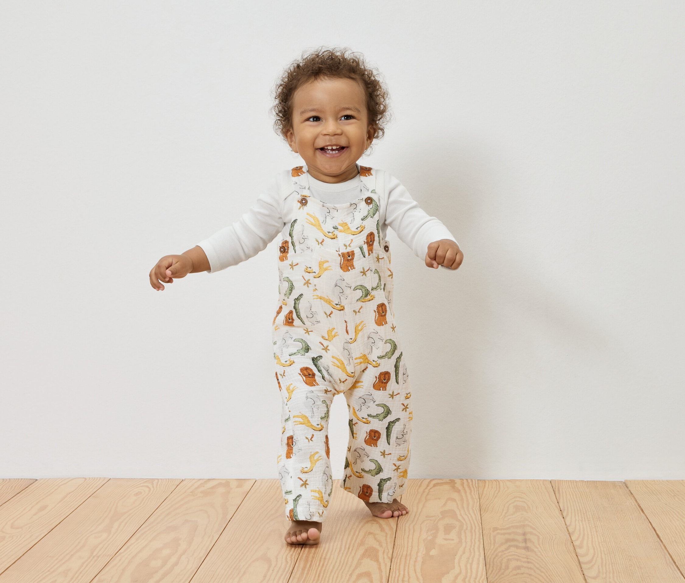 Uśmiechnięte dziecko z kręconymi włosami stoi na drewnianej podłodze, ubrane w białą bluzkę z długim rękawem pod białym kombinezonem w zwierzęcy wzór.