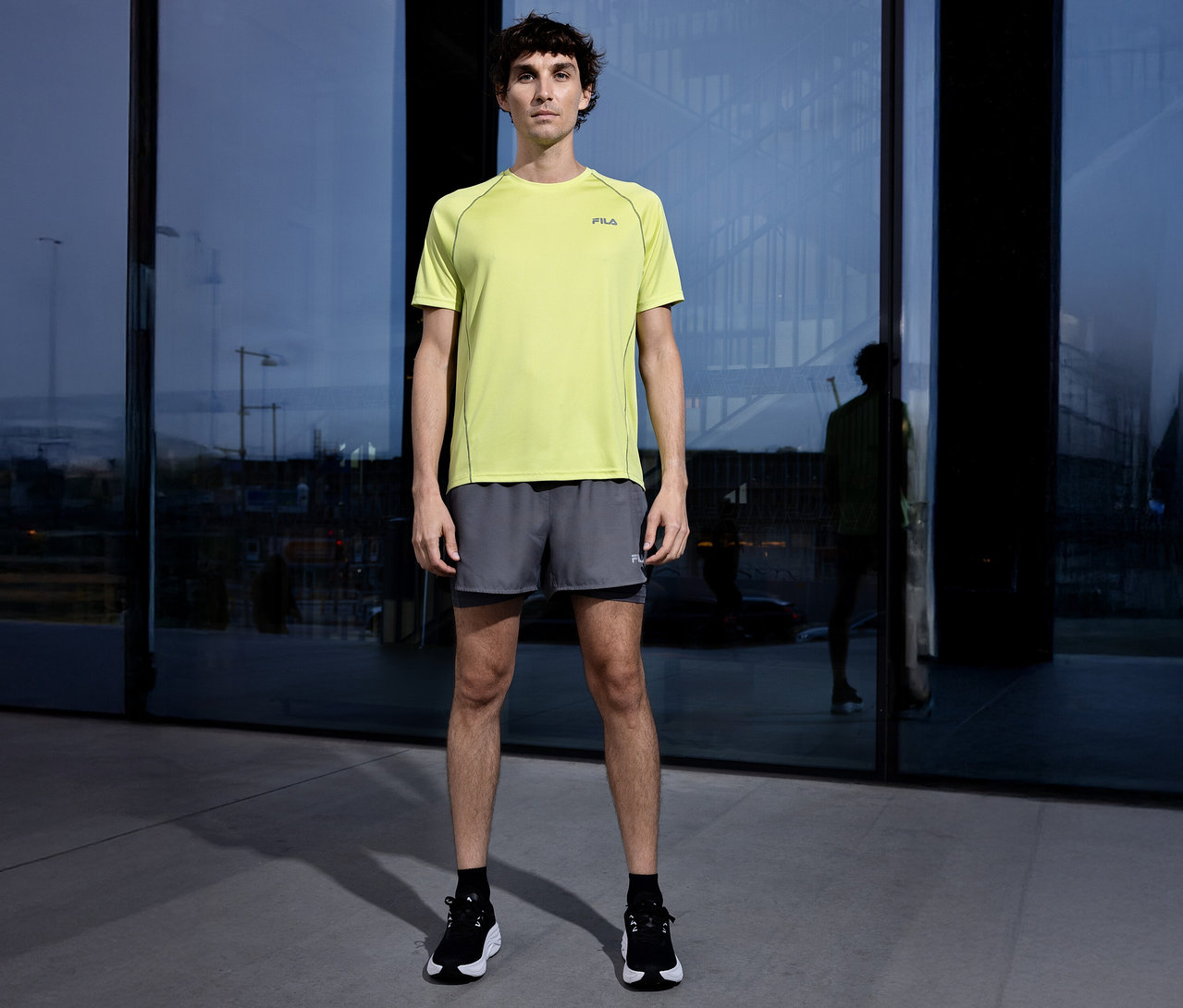 Mężczyzna stoi przed budynkiem ubrany w żółtą koszulkę Fila, szare szorty i czarne buty.