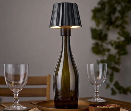 Lampa stołowa w kształcie butelki wina stoi na drewnianym stole obok dwóch kieliszków do wina i miski orzechów.