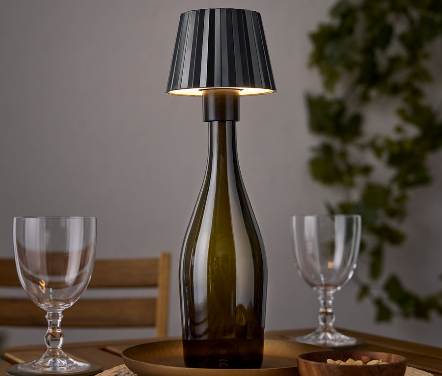 Lampa stołowa w kształcie butelki wina stoi na drewnianym stole obok dwóch kieliszków do wina i miski orzechów.