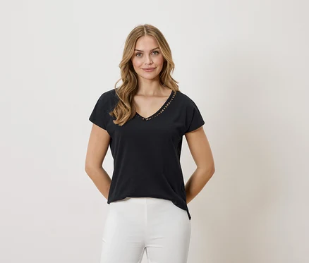 Kobieta o blond włosach ma na sobie czarny T-shirt z dekoltem w serek i białe spodnie na neutralnym tle.