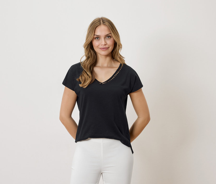 Kobieta o blond włosach ma na sobie czarny T-shirt z dekoltem w serek i białe spodnie na neutralnym tle.