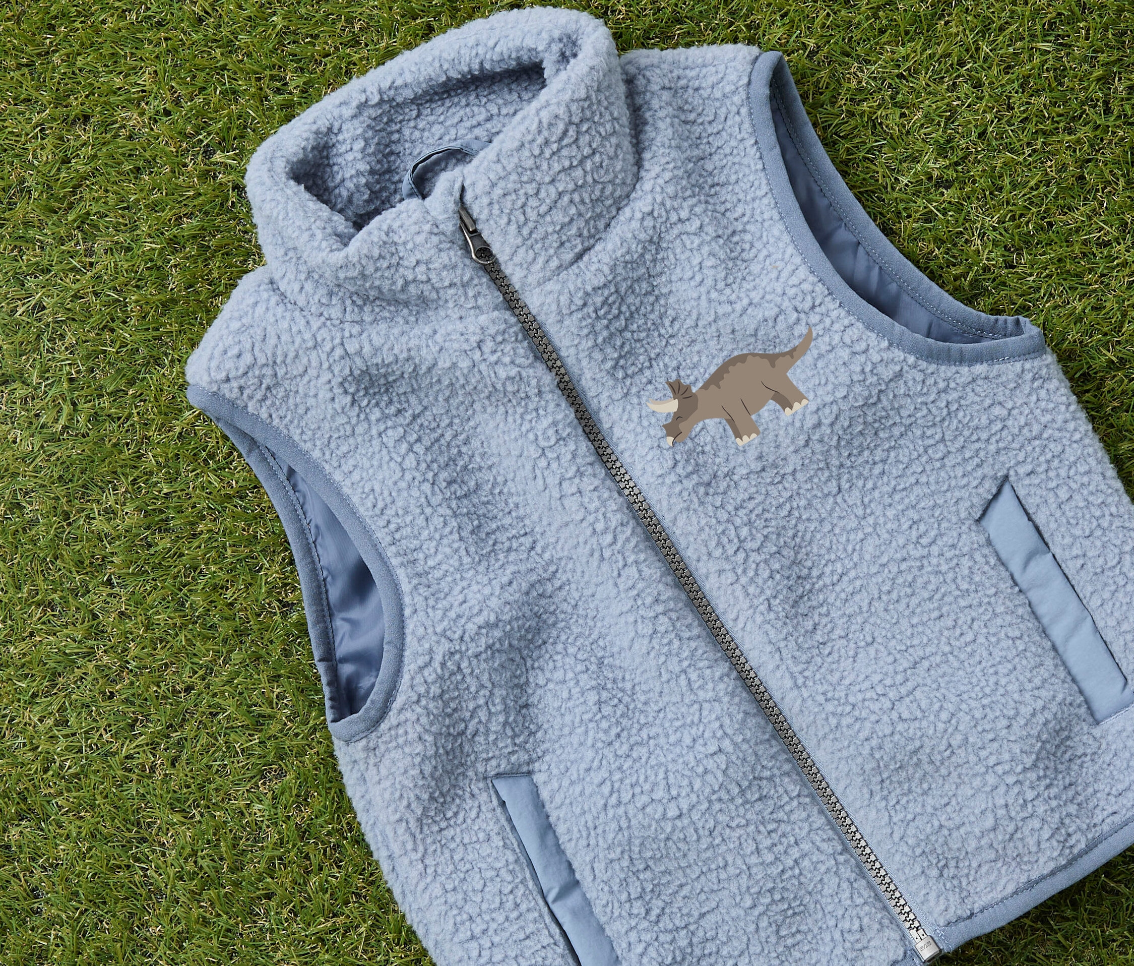 Jasnoniebieska kamizelka polarowa z motywem triceratopsa leży na zielonej trawie.