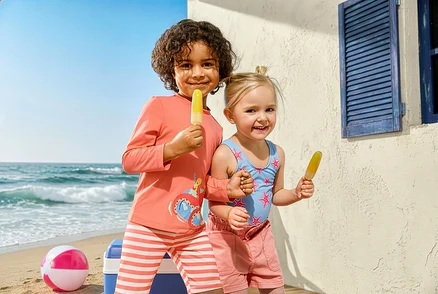 Dwoje dzieci z lodami na plaży. Dziewczynka ma na sobie strój kąpielowy Kinder-Badeanzug, Seesterne i dżinsowe szorty. Chłopiec nosi kombinezon Kinder-Kombi z ochroną UV i motywem ośmiornicy.