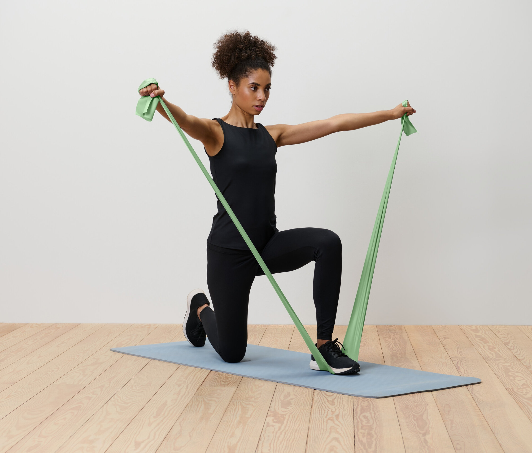 Kobieta klęczy na niebieskiej macie do jogi, rozciągając zieloną gumę oporową.