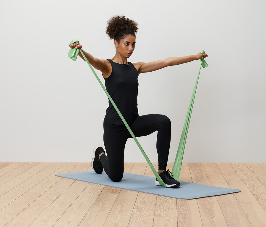 Kobieta klęczy na niebieskiej macie do jogi, rozciągając zieloną gumę oporową.