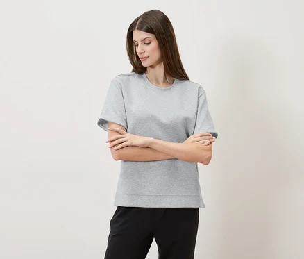 Kobieta ze skrzyżowanymi ramionami ma na sobie szary T-shirt i czarne spodnie na neutralnym tle.