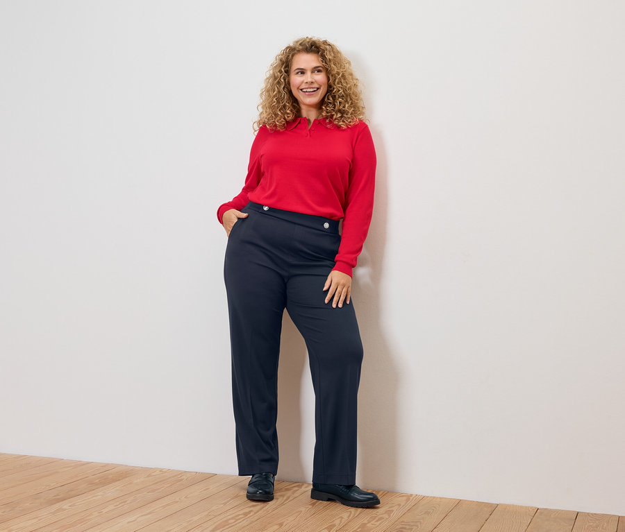 Kobieta z kręconymi włosami, w czerwonej bluzce i niebieskich spodniach.