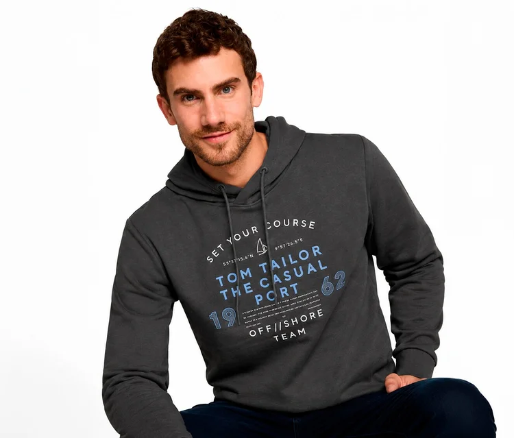 Uśmiechnięty mężczyzna w ciemnoszarej bluzie z kapturem z grafiką „TOM TAILOR THE CASUAL PORT” i żaglówką. Nosi ciemnoniebieskie dżinsy.