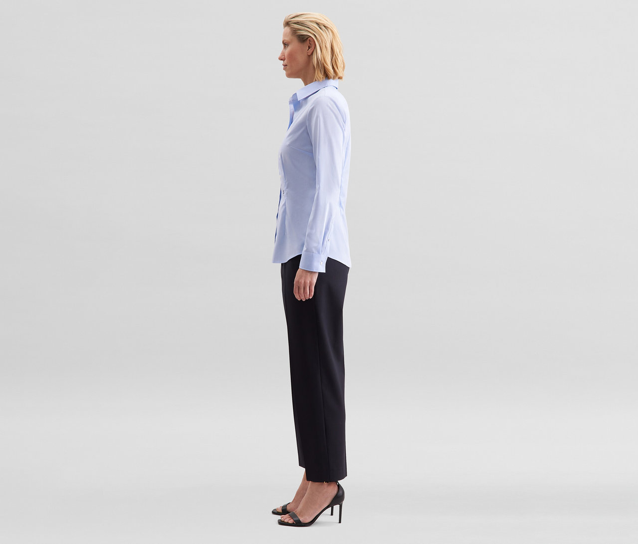Kobieta stoi profilem, ubrana w jasnoniebieską bluzkę Seidensticker Slim Fit non iron, czarne spodnie i czarne sandały na obcasie.