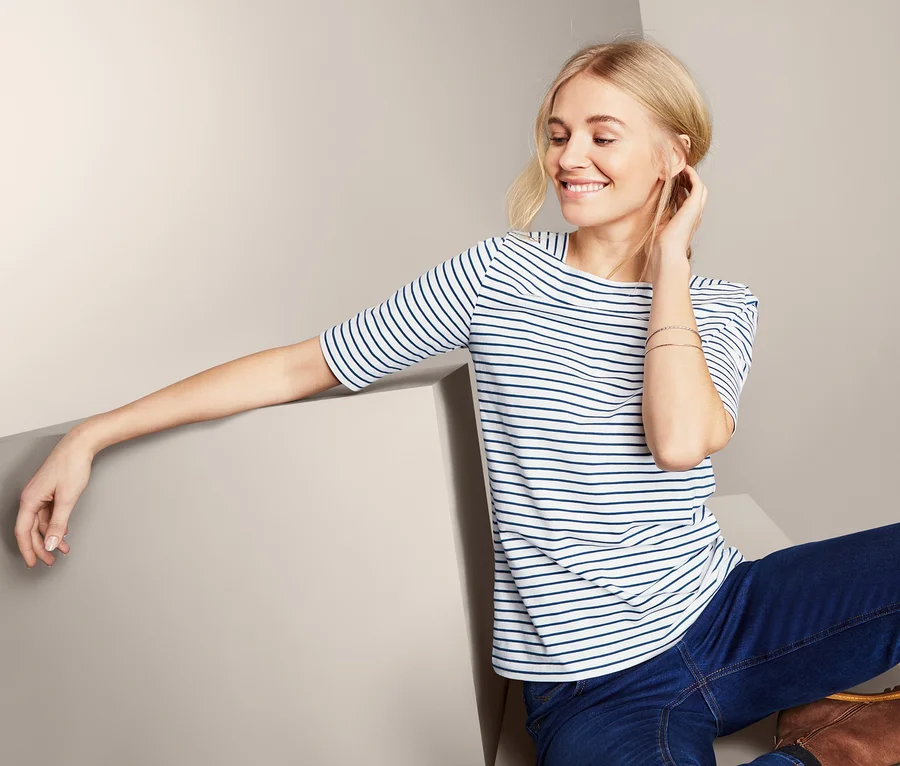 Blondynka siedzi i ma na sobie damską bluzkę z półdługimi rękawami w paski, dżinsy i brązowe buty.