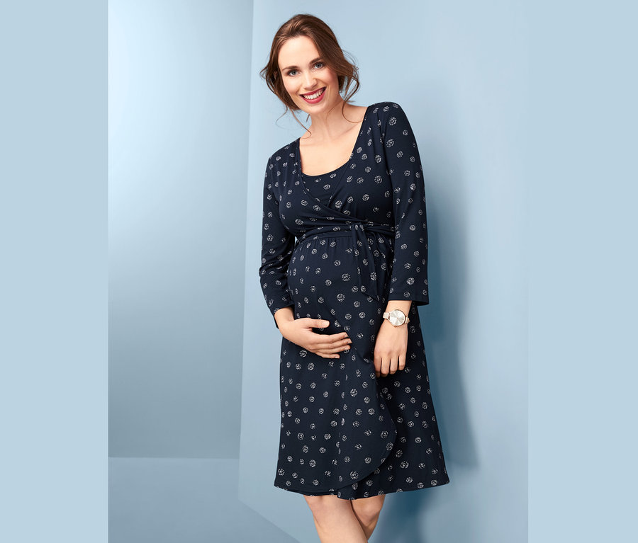 Uśmiechnięta kobieta w ciąży trzyma się za brzuch, ubrana w granatową sukienkę ciążową stylizowaną na kopertową, z nadrukiem.