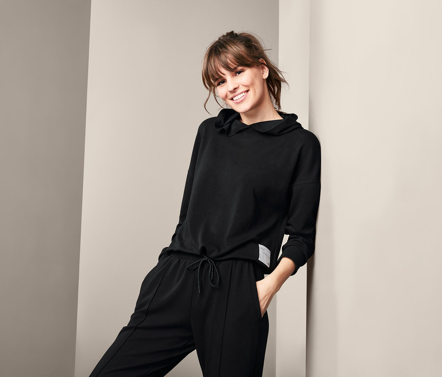 Uśmiechnięta kobieta opiera się o ścianę w czarnej damskiej bluzie rekreacyjnej z dzianiny dresowej i spodniach.