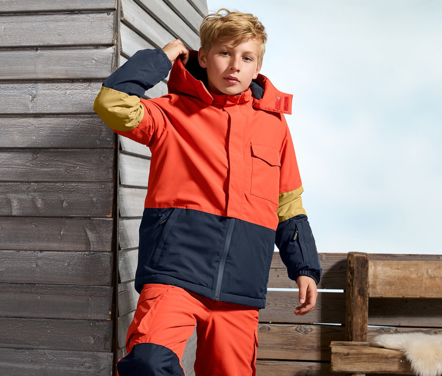 Chłopiec w pomarańczowo-granatowej kurtce snowboardowej.