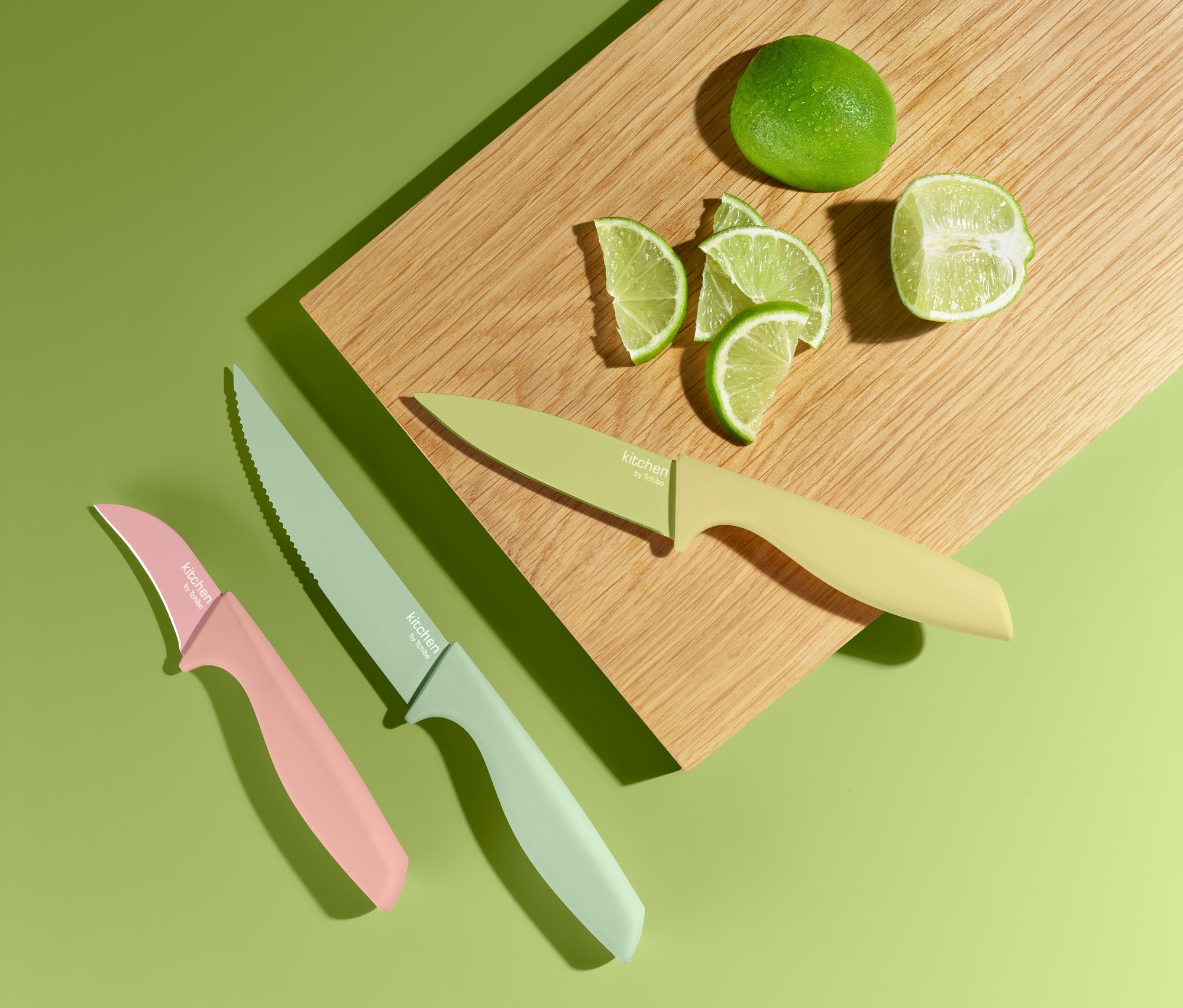 Na zielonym tle leży drewniana deska z limonką i pokrojonymi kawałkami limonki obok zestawu 3 noży kuchennych.