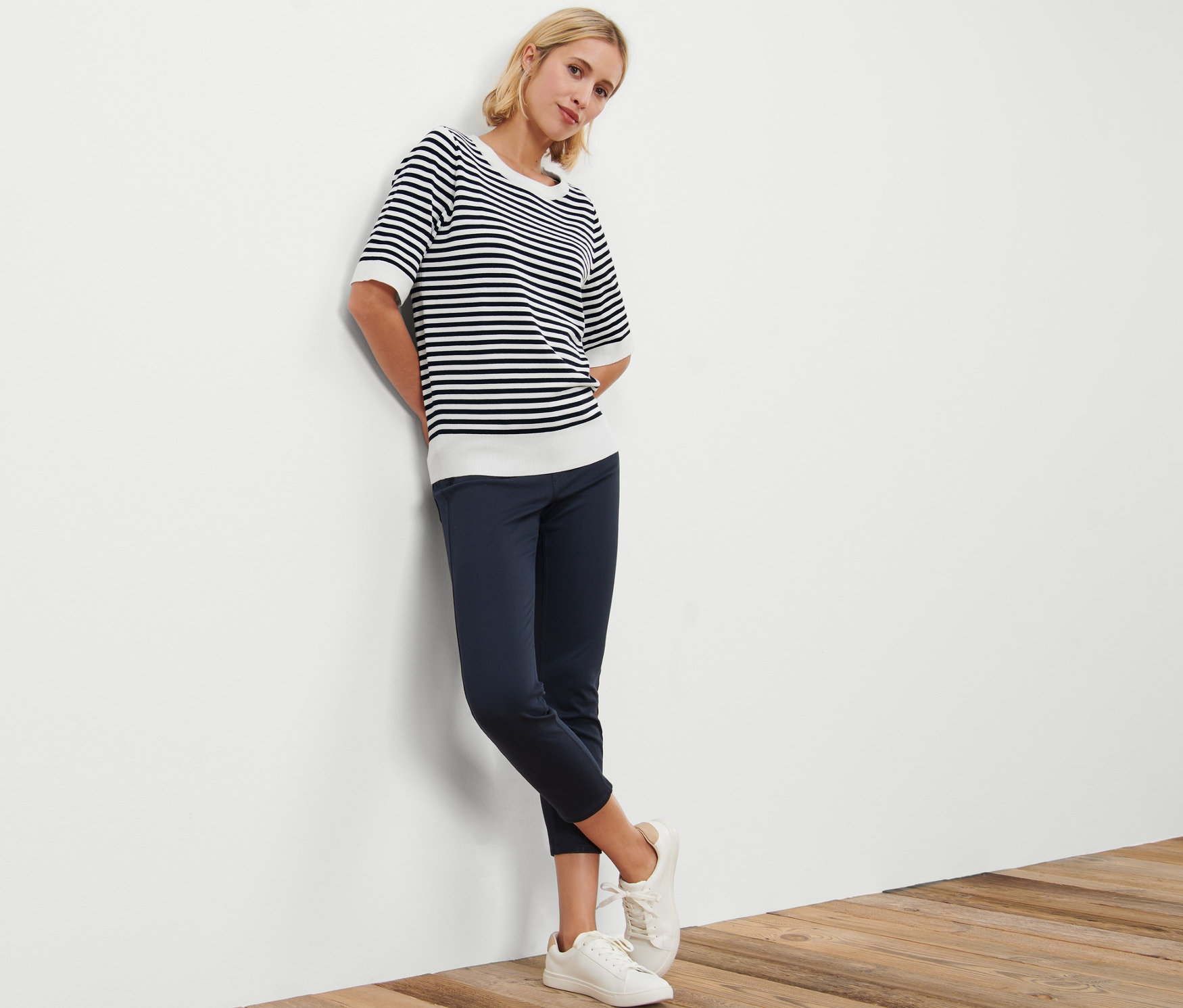 Kobieta oparta o ścianę ma na sobie sweter w paski z cienkiej dzianiny, granatowe spodnie i białe tenisówki.