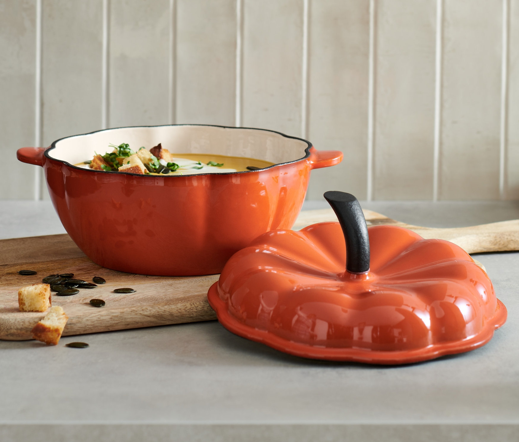 Otwarty żeliwny garnek w kształcie dyni wypełniony zupą z grzankami stoi na drewnianej desce obok pokrywki.