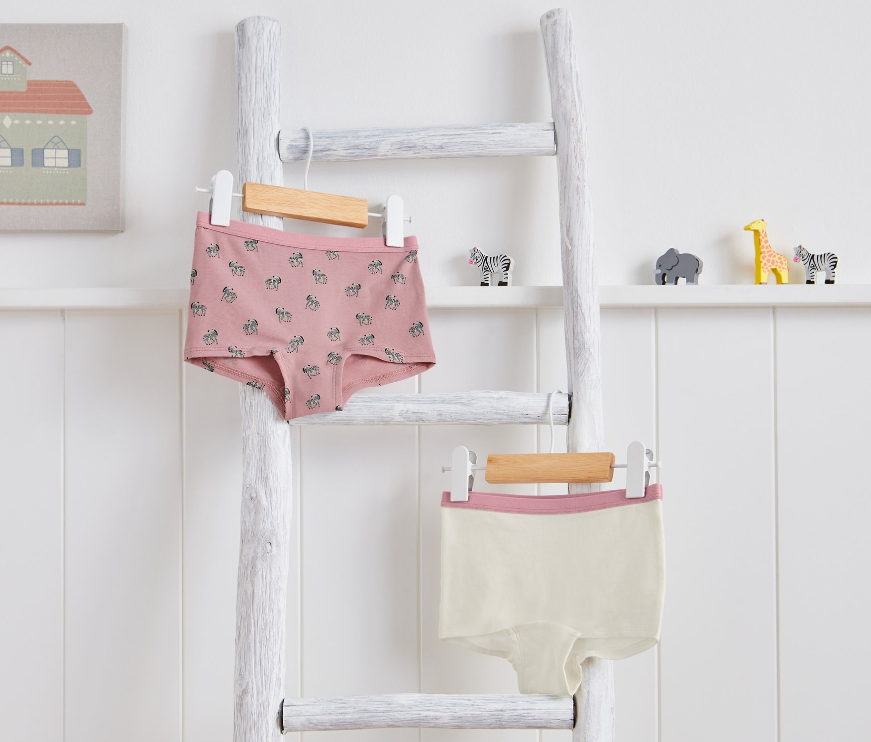 Figi dziecięce, 2 pary, wiszą na białej drabinie. Jedne są różowe we wzór zebry, drugie kremowe z różowym brzegiem.