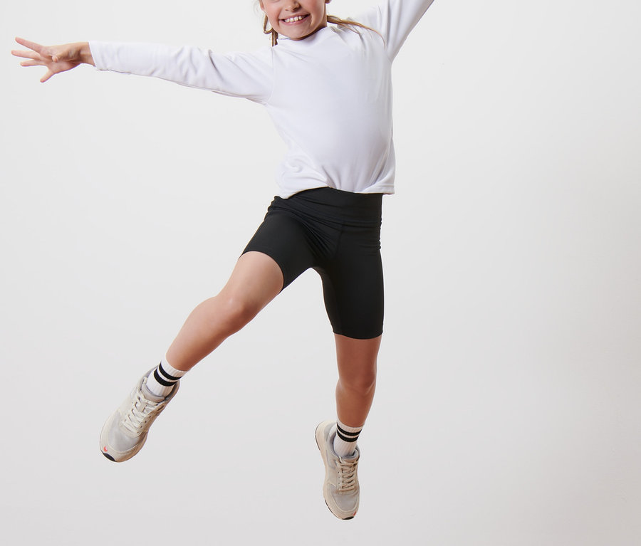 Dziewczynka skacze na drewnianym podeście, ubrana w czarne krótkie legginsy i białą bluzkę z długim rękawem. Ma na sobie Kolarki, 2 pary.