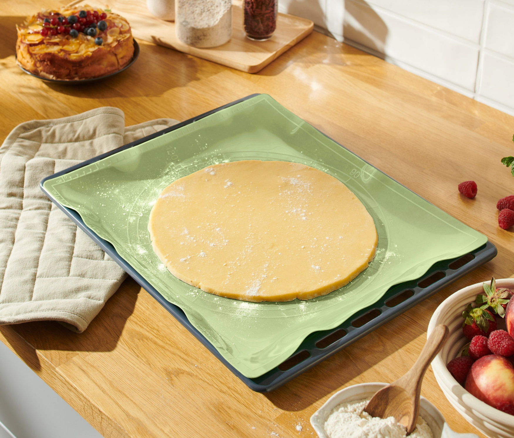 Na blacie leży wielorazowa mata do pieczenia z rozwałkowanym ciastem, obok leży rękawica kuchenna i miska z owocami.