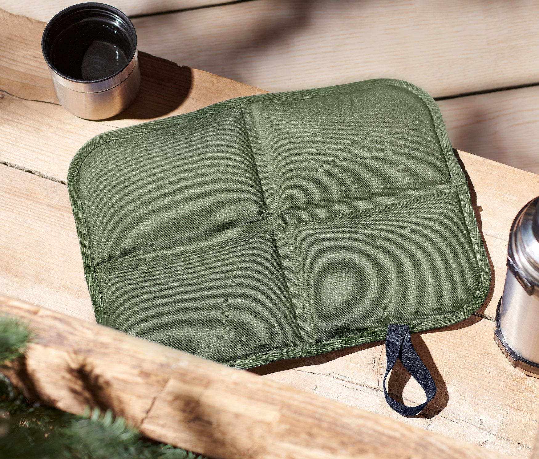 Na drewnianym stole leżą: zielona składana poduszka termiczna do siedzenia, termos i kubek.