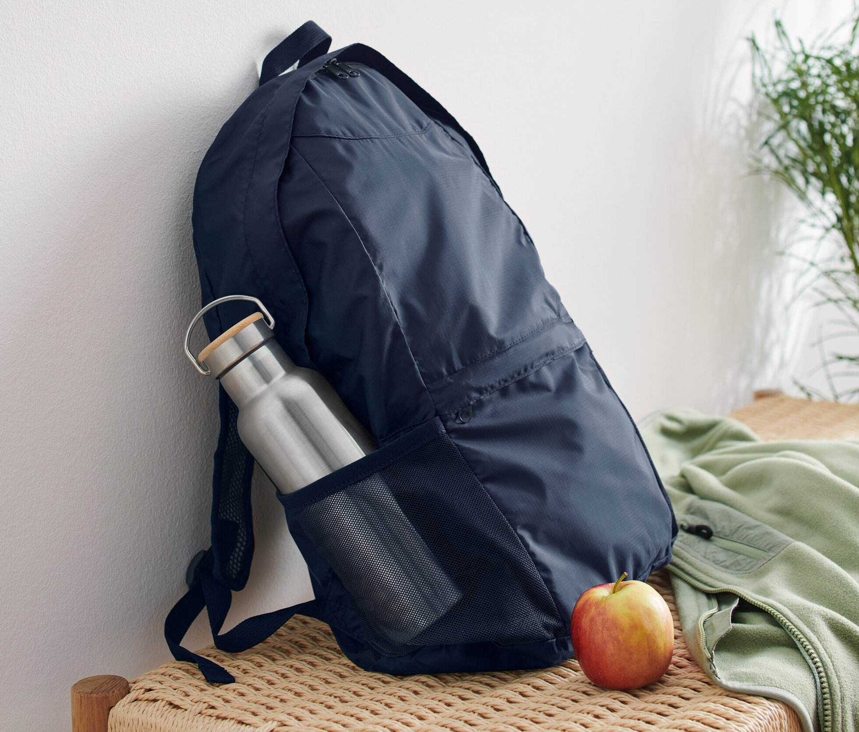 Granatowy składany plecak oparty o białą ścianę na wiklinowej ławce. W bocznej kieszeni plecaka znajduje się srebrna butelka na wodę, a obok plecaka leży jabłko.