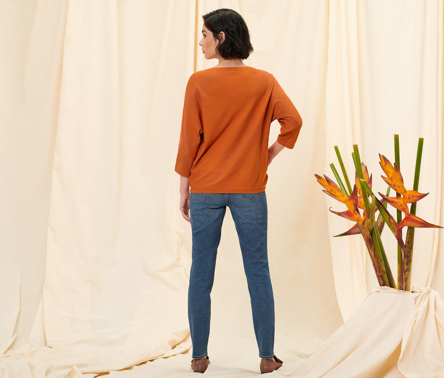 Kobieta stoi tyłem do aparatu, ubrana w jeansy slim »Fit Emma« mid blue denim, pomarańczowy sweter z delikatnej dzianiny i półbuty ze skóry welurowej.