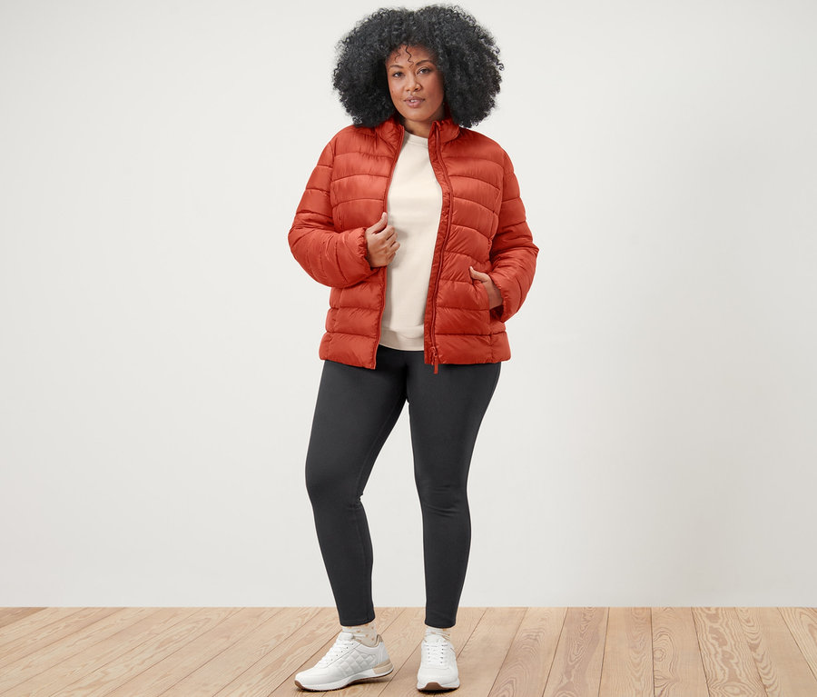 Kobieta pozuje w pomarańczowej pikowanej kurtce, bluzie z haftem i jegginsach.