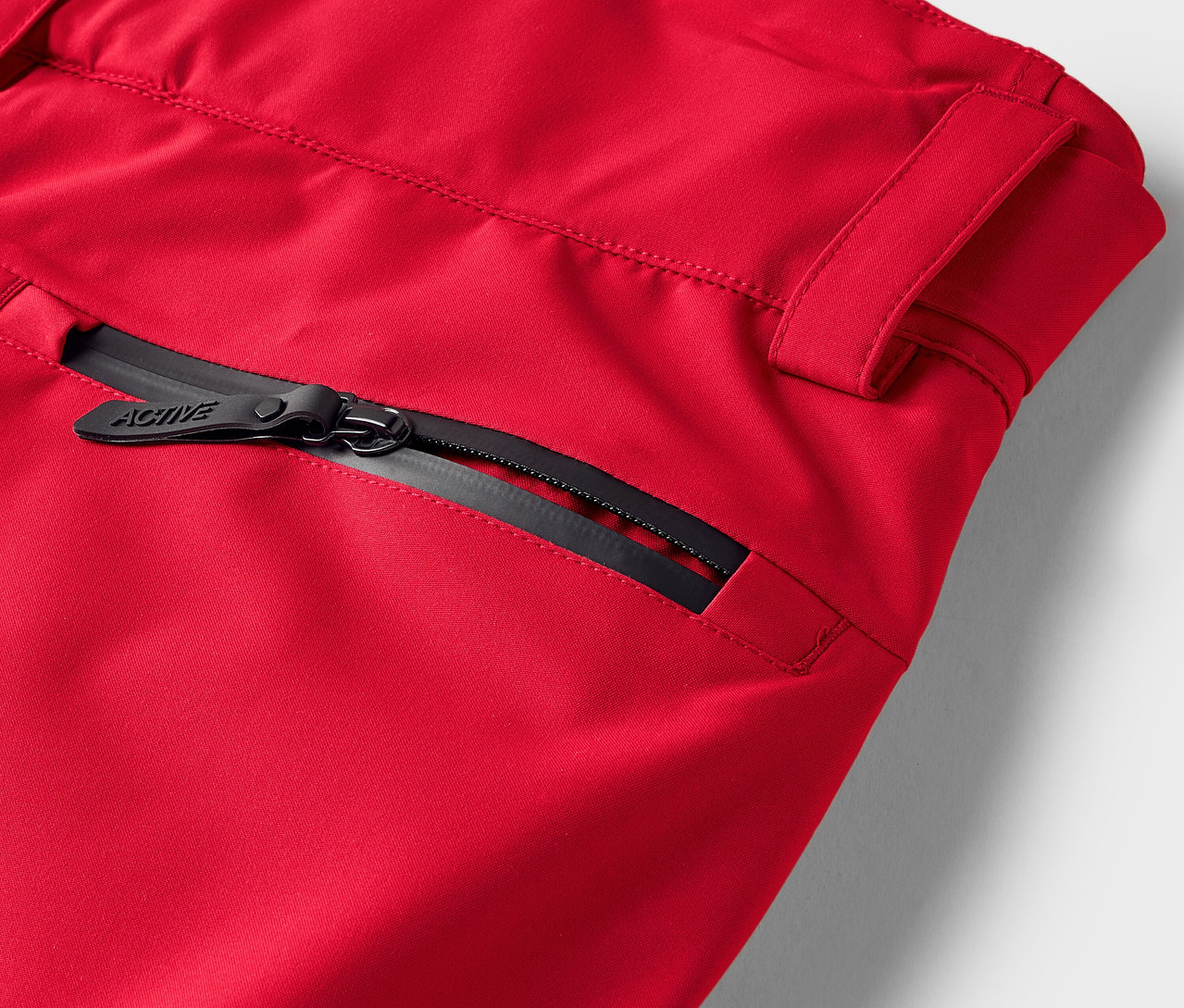 Detal czerwonych spodni z czarnym zamkiem błyskawicznym na kieszeni.