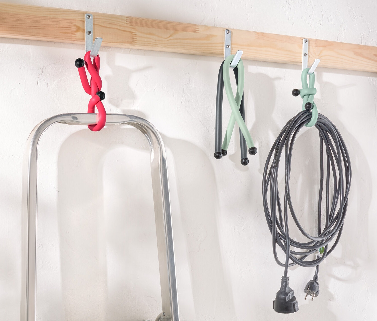 Drabina stoi pod drewnianą listwą z haczykami, na których wiszą Power Kabelbinder w kolorze czerwonym, zielonym i czarnym.